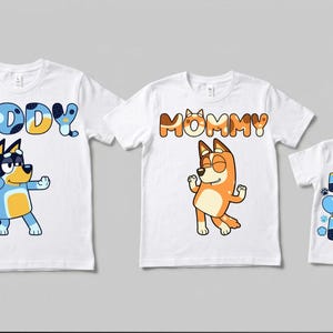 Puede incluir: Tres camisetas blancas con personajes de perros de dibujos animados. La primera camiseta dice DADDY con un perro azul. La segunda camiseta dice MOMMY con un perro naranja. La tercera camiseta dice BIRTHDAY BOY con un perro azul y huellas.