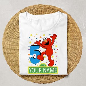 Könnte beinhalten: Ein weißes T-Shirt mit einer Grafik der roten Figur Elmo, einer großen blauen Zahl 5 sowie bunten Konfettipunkten und Sternen. Unter der Figur befindet sich ein gelbes rechteckiges Schild mit dem Text 123 und dem Platzhaltertext YOUR NAME.