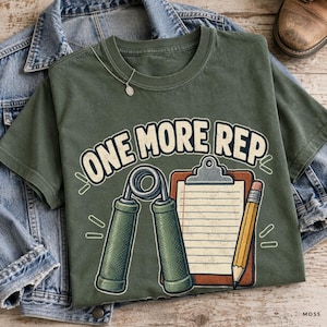 May include: An olive green t-shirt featuring a graphic print with the text ONE MORE REP in bold letters. The design includes a metal hand grip strengthener, a brown clipboard with lined paper, and a yellow pencil. A silver necklace rests on the collar.