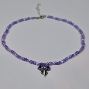 Könnte beinhalten: Eine Perlenkette mit abwechselnd kleinen violetten und weißen Perlen in kreisförmiger Anordnung. Ein silberfarbener Metallanhänger in Form einer Schleife mit zwei dunkelvioletten herzförmigen Akzenten hängt in der Mitte der Kette.