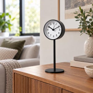 May include: A black analog table clock with a round face, white dial, and black numerals. The clock is mounted on a tall, slender black metal stand with a circular base, featuring black hour, minute, and second hands on a clean white background.