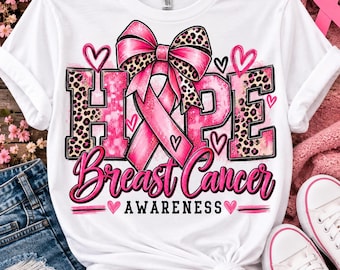 May include: A white t-shirt featuring a graphic design with the word HOPE in large block letters with a pink and leopard print pattern. A pink awareness ribbon with a leopard print bow sits above the text, accompanied by pink hearts and the text Breast Cancer AWARENESS.