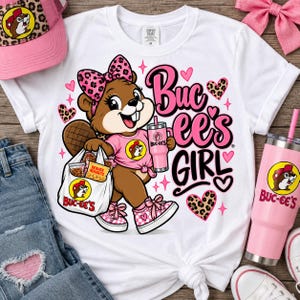May include: A white t-shirt featuring a cartoon beaver wearing a pink leopard print bow, a pink shirt, and pink trainers. The beaver holds a shopping bag and a pink tumbler. The text reads Buc-ee's GIRL, surrounded by pink and leopard print hearts.