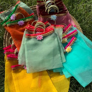 Può includere: Una collezione di sacchetti regalo in organza trasparente in vari colori tra cui giallo, arancione, verde salvia, verde acqua e bordeaux. Ogni sacchetto presenta un bordo dorato, un manico circolare dorato e un fiore di loto.