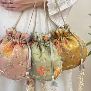 May include: Three drawstring pouches in dusty pink, sage green, and mustard yellow, each featuring a floral print. The bags have metallic braided handles, beaded trim along the top edge, and decorative tassels with hanging pearl-like beads.