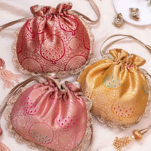 May include: Three drawstring pouches featuring intricate metallic brocade patterns in red, gold, and pink. Each bag has a scalloped edge with beaded trim and long braided cords ending in beaded tassels. The text RITU KUMAR HOME is visible.