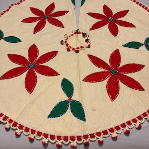 May include: A circular off-white fabric tree skirt featuring red felt poinsettia appliqués with teal beaded centers and green felt leaf accents. The edges are finished with a red and white crocheted trim featuring small red pom-pom details.