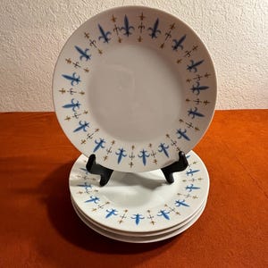 May include: A stack of four white ceramic dinner plates with a blue and gold fleur-de-lis pattern around the rim.