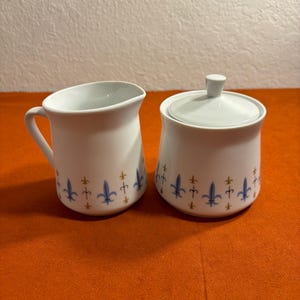May include: White ceramic creamer and sugar bowl set. The creamer has a handle and spout. Both pieces are decorated with blue and gold fleur-de-lis designs. The sugar bowl has a lid with a small knob.