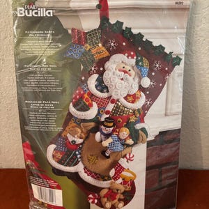 May include: A Plaid Bucilla Patchwork Santa felt stocking kit in original packaging. The design features a Santa Claus figure, a snowman, a doll, and a teddy bear. The stocking is decorated with sequins, beads, and patchwork patterns, measuring 18 inches.