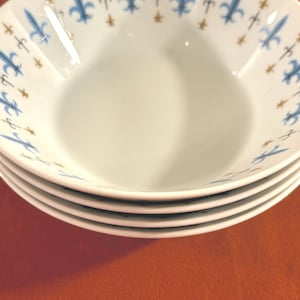 May include: A stack of four white ceramic bowls with a blue and gold fleur-de-lis pattern around the rim.