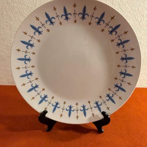 May include: A white ceramic plate with a blue and gold fleur-de-lis design around the rim. The plate is on a black stand.
