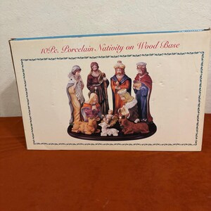 May include: A rectangular product box for a 10 Pc. Porcelain Nativity on Wood Base. The box features a printed illustration of a ten-piece porcelain nativity set, including figures of people and animals arranged on a dark oval wooden base.