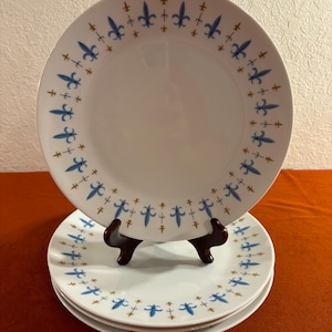 May include: Set of four white ceramic dinner plates with a blue and gold fleur-de-lis pattern around the rim.