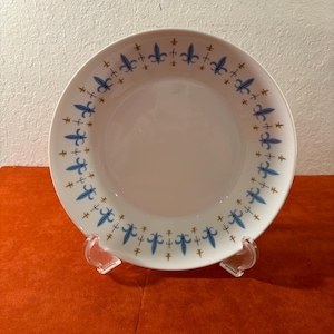 May include: A white ceramic bowl with a blue and gold floral pattern around the rim. The bowl is on a clear plastic stand.
