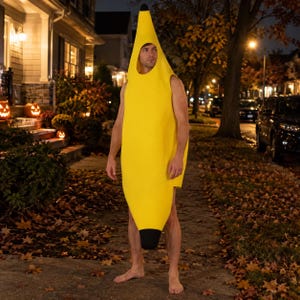 Op de afbeelding: Een felgeel bananenkostuum met een gebogen, langwerpige lichaamsvorm, een puntige bovenkant en een afgeronde onderkant. Het kostuum is voorzien van een zwarte punt aan zowel de boven- als onderkant en een grote ronde gezichtsopening.