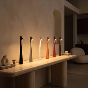 Op de afbeelding: Een rij van zes slanke, conische tafellampen in zwart, lichtgrijs, wit, oranje, rood en bruin. Elke lamp heeft een smalle voet die naar boven toe taps toeloopt naar een kleine, schuine lichtbron die een warme gloed op de muur werpt.