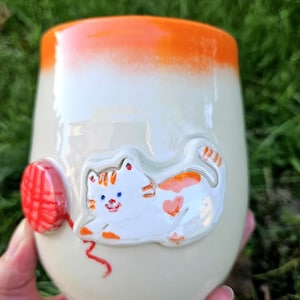 May include: A ceramic tumbler with a glossy off-white finish and a bright orange rim. The exterior features a raised, hand-painted white cat with orange tabby markings and a heart-shaped spot, playing with a textured red ball of yarn.