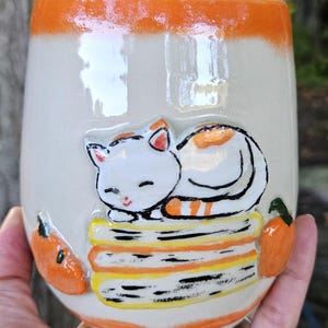 May include: A ceramic mug with a glossy off-white finish and a bright orange rim. The front features a raised, hand-painted design of a white cat with orange spots sleeping on a stack of three books, flanked by two small orange pumpkins.