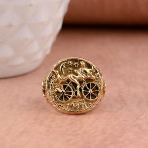 Puede incluir: Un anillo de tono dorado con una cara circular que presenta un diseño en relieve de una figura montando una bicicleta bajo una luna creciente y estrellas. La parte inferior de la cara del anillo muestra el año 1943 grabado, con detalles intrincados.