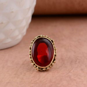 May include: A ring featuring a large, oval-shaped, deep red gemstone set in a gold-toned metal frame. The setting is characterised by a decorative, scalloped edge with small, rounded metal beads encircling the central stone in a detailed pattern.