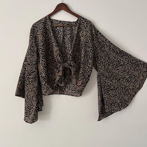 Könnte beinhalten: Eine bauchfreie Bluse mit tiefem V-Ausschnitt und einer Bindung vorne. Das Kleidungsstück besteht aus einem leichten Stoff mit einem schwarzen und beigen abstrakten Tiermuster. Es hat lange, weite Glockenärmel, die sich ab den Schultern ausstellen.