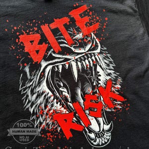 Op de afbeelding: Een zwart t-shirt met een witte grafische illustratie van een grommende wolf met een open bek en scherpe tanden. De tekst BITE RISK is in dikke rode letters over de wolf gedrukt, vergezeld van rode verfspatten als effect.
