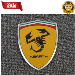 Könnte beinhalten: Ein schildförmiges Abarth-Emblem mit gelbem Hintergrund und einer schwarzen Skorpion-Grafik in der Mitte. Der obere Teil des Schildes zeigt horizontale Streifen in Grün, Weiß und Rot. Das Wort ABARTH steht darunter in schwarzen Großbuchstaben.