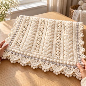 May include: A cream-colored crocheted blanket featuring vertical cable stitch patterns, intricate openwork lace sections, and a decorative border with small bobbles and scalloped edges accented by a thin line of grey yarn.