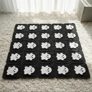 May include: A square crochet blanket composed of twenty-five individual black squares, each featuring a white ghost motif with two small black eyes and a curved mouth. The blanket is arranged in a five-by-five grid pattern with uniform stitching.