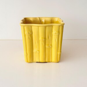 May include: A square-shaped ceramic planter with a glossy yellow finish. The exterior features a molded relief pattern that mimics the appearance of vertical bamboo stalks, complete with horizontal nodes and segments around the entire perimeter.