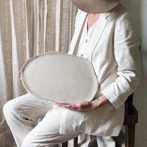 Può includere: Un grande vassoio in ceramica color bianco sporco con una forma ovale organica e irregolare e una finitura liscia e opaca. Il vassoio presenta bordi leggermente rialzati e irregolari e una superficie ampia e piatta, dall'aspetto neutro e semplice.