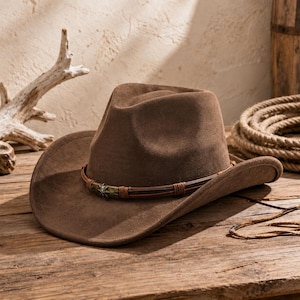 Può includere: Un cappello da cowboy in feltro marrone con corona pizzicata e tesa larga e curva. Il cappello è decorato con una fascia composta da sottili cordini in pelle marrone e una fibbia metallica centrale con un emblema a forma di stella.