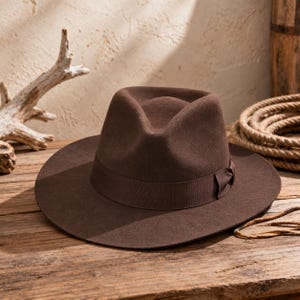 May include: A dark brown felt fedora hat featuring a structured crown with a centre crease and a wide, flat brim. The hat is encircled by a matching dark brown grosgrain ribbon band that includes a small, folded bow detail on the side.