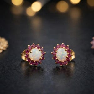 May include: A pair of gold stud earrings featuring a central round white opal cabochon surrounded by a halo of small, faceted, deep pink rubies. The stones are set in a scalloped metal frame, creating a floral-inspired cluster design.