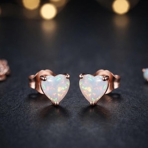 Könnte beinhalten: Ein Paar herzförmige Ohrstecker mit weißen, opalisierenden Steinen und irisierenden Sprenkeln, gefasst in roségoldfarbenem Metall. Die Ohrringe haben einen klassischen Steckverschluss und eine glatte, polierte Oberfläche.
