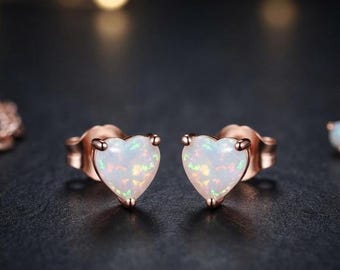 Könnte beinhalten: Ein Paar herzförmige Ohrstecker mit weißen, opalisierenden Steinen und irisierenden Sprenkeln, gefasst in roségoldfarbenem Metall. Die Ohrringe haben einen klassischen Steckverschluss und eine glatte, polierte Oberfläche.