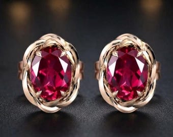 May include: A pair of earrings featuring large, oval-cut, deep red gemstones set in a polished, twisted gold-tone metal frame. The design showcases a central faceted ruby-colored stone surrounded by a continuous, interwoven metallic border.