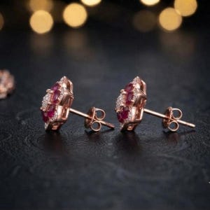 May include: A pair of rose gold stud earrings featuring a central cluster of deep red rubies surrounded by a halo of small white diamonds. The earrings have a secure post back mechanism and are designed with a floral-inspired arrangement of stones.