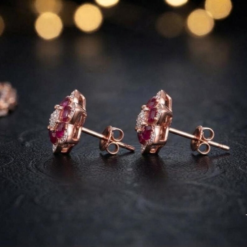 May include: A pair of rose gold stud earrings featuring a central cluster of deep red rubies surrounded by a halo of small white diamonds. The earrings have a secure post back mechanism and are designed with a floral-inspired arrangement of stones.
