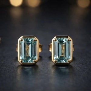 Könnte beinhalten: Ein Paar Ohrstecker mit rechteckigen, im Smaragdschliff geschliffenen hellblauen Edelsteinen in goldfarbenen Metallfassungen. Jeder Ohrstecker ist mit vier kleinen Krappen versehen, die den Stein sichern und ein klares, symmetrisches Design bilden.