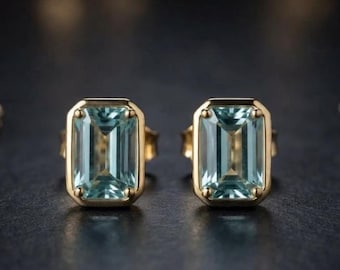May include: A pair of stud earrings featuring rectangular emerald-cut light blue gemstones set in gold metal frames. Each earring has four small prongs securing the stone, creating a clean and symmetrical design with a polished metallic finish.
