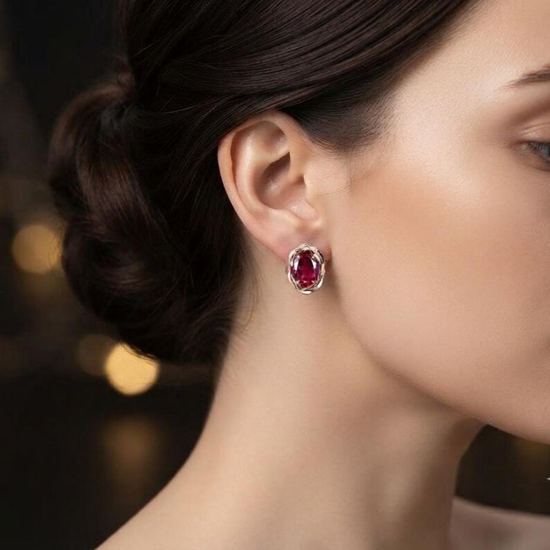 Peut inclure: Une boucle d'oreille orn&eacute;e d'une pierre pr&eacute;cieuse rouge fonc&eacute; de forme ovale, mont&eacute;e sur une monture en m&eacute;tal argent&eacute; poli. Le sertissage pr&eacute;sente un motif d&eacute;licat en fil torsad&eacute; qui entoure la pierre centrale, maintenue par des griffes.