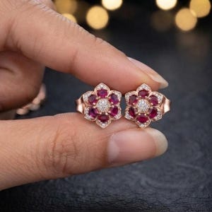May include: A pair of rose gold stud earrings shaped like flowers, each featuring six deep red oval-cut gemstones arranged as petals around a central cluster of small, clear white round-cut stones set in a metallic frame.