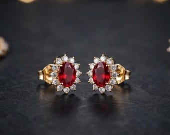 Può includere: Un paio di orecchini a lobo in oro con una pietra rossa centrale dal taglio ovale circondata da un alone di piccole pietre bianche rotonde. Gli orecchini presentano un classico design floreale con una montatura in metallo dorato lucido.