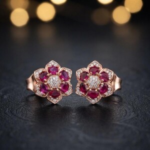 May include: A pair of rose gold stud earrings shaped like flowers. Each earring features six oval-cut deep red rubies arranged as petals around a central cluster of small, round, clear white diamonds, with additional diamonds set along the edges.