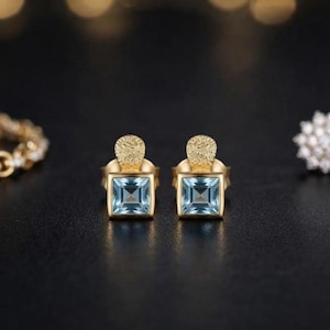Könnte beinhalten: Ein Paar Ohrstecker aus Gold mit hellblauen Edelsteinen im Quadratschliff. Jeder Ohrstecker verfügt über ein strukturiertes, halbkreisförmiges Goldoberteil, das an der quadratischen Fassung befestigt ist und einen facettierten blauen Stein zeigt.