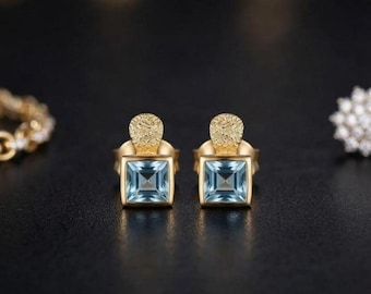 May include: A pair of gold stud earrings featuring square-cut light blue gemstones. Each earring has a textured, semi-circular gold top piece attached to the square setting, showcasing a clear, faceted blue stone in a polished gold frame.