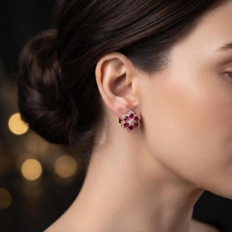 May include: A floral-shaped stud earring featuring six round, deep red rubies arranged in a circle around a central clear diamond, all set in a polished gold metal frame, worn on the earlobe with a secure backing mechanism.