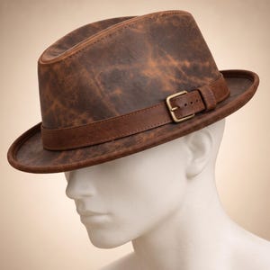 Pode incluir: Um chapéu fedora castanho com uma textura desgastada e manchada, apresentando uma faixa de couro castanho escuro com uma fivela de metal em tom de latão. O chapéu tem uma copa estruturada e uma aba curva, exibindo uma aparência envelhecida.